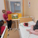 W sali szpitalnej dziewczynka i chłopiec siedzą na swoich łóżkach, a nauczycielka siada przy stoliku, na którym ustawiony jest teatr kamishibai.