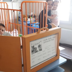 W sali szpitalnej chłopiec siedzący na łóżku i mama z dzieckiem na ręku oglądają teatr kamishibai ustawiony na szafce.