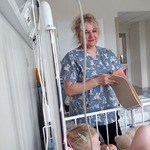 Nauczycielka jest przy łóżku dziecka i zachęca je do wybrania kolorowej kartki do wykonania laurki