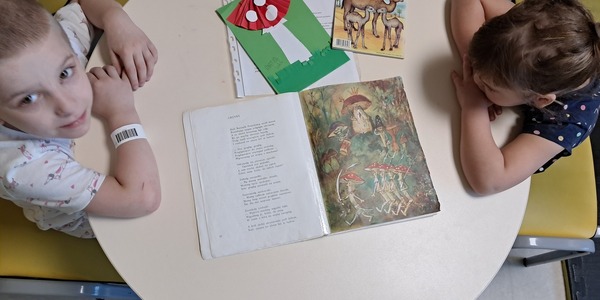 Dwoje dzieci siedzi przy stoliku, na którym leży książka z tekstem wiersza.