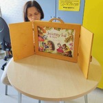 Nauczycielka siedząca przy stoliku w świetlicy oddziału Neurologii w UDSK z teatrzykiem kamishibai i widoczną w nim stroną tytułową bajki „Jesienny bal w lesie”.