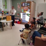 Świetlica oddziału Pediatrii i Neurologii  w WSZ wypełniona siedzącymi pacjentami i rodzicami oglądającymi przedstawienie i nauczycielka stojąca obok stolika ze skrzynką teatrzyku kamishibai .