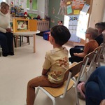 Mali pacjenci siedzący w świetlicy oddziału Pediatrii i Neurologii  w WSZ i oglądający bajkę prezentowaną przez nauczycielkę siedzącą przy stoliku z teatrzykiem kamishibai.