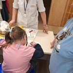 Dziewczynka siedzi przy stole i wykonuje ćwiczenie „zbieranie kropelek”, a nauczycielka stoi i obserwuje to.