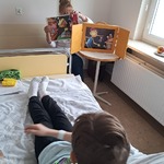 Chłopiec leży w łóżku szpitalnym, a nauczycielka zmienia planszę w teatrze kamishibai.