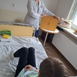 Nauczycielka na sali prezentuje dziecku leżącemu w łóżku skrzynkę butai teatru kamishibai.