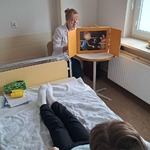 Nauczycielka czyta bajkę w teatrze kamishibai chłopcu leżącemy w łóżku szpitalnym.