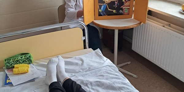 Nauczycielka czyta bajkę w teatrze kamishibai chłopcu leżącemy w łóżku szpitalnym.