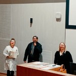 Po lewej nauczycielka trzyma podziękowanie, po środku mężczyzna mówi do mikrofonu, po prawej stoi dyrektor szkoły