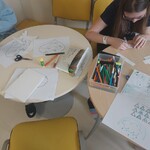 dziewczyna przy stole rysuje na drugim stole stoją kredki i leżą kartki z zadaniami i nożyczki