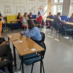 sala szkolna przy stolikach siedzi młodzież i nauczyciele przy jednym ze stołów stoi kobieta prowadząca szkolenie