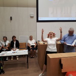 stół, za stołem siedzą trzy kobiety obok podium kobieta z podniesionymi rękami i mężczyzna z książką