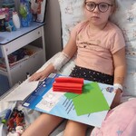 Dziewczynka siedzi w łóżku i wykonuje pracę plastyczną.