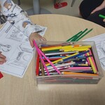 Na stoliku stoi pudełko kredek, leżą karty pracy i widoczne są ręce dzieci 
wykonujących zadania na tych kartach.