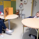 W świetlicy szpitalnej nauczycielka siedzi przy stoliku i przedstawia bajkę w teatrzyku kamishibai, a troje dzieci ogląda przedstawienie.