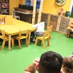 Nauczycielka siedzi przy żółtych stolikach w świetlicy szpitalnej i czyta bajkę w teatrzyku kamishibai, a mali pacjenci z rodzicami słuchają i oglądają przedstawienie.