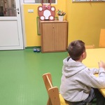 Nauczycielka prezentująca bajkę w teatrzyku kamishibai i chłopiec w szarej                           bluzie siedzą przy żółtych stolikach w świetlicy, a dzieci i rodzice siedzą na                           pufach i oglądają przedstawienie.