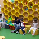 Mali pacjenci i rodzice siedzą na żółtych pufach i krzesełkach na tle ściany,  na której wiszą półki w kształcie plastra miodu.
